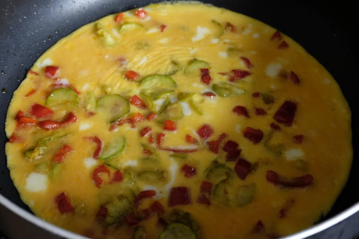 Eggs poured over cooked zucchini and red bell pepper in a frying pan