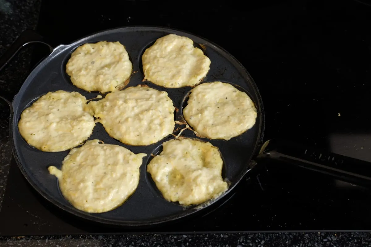 Small portions of potato and zucchini batter cooking on their first side in a pancake-well pan
